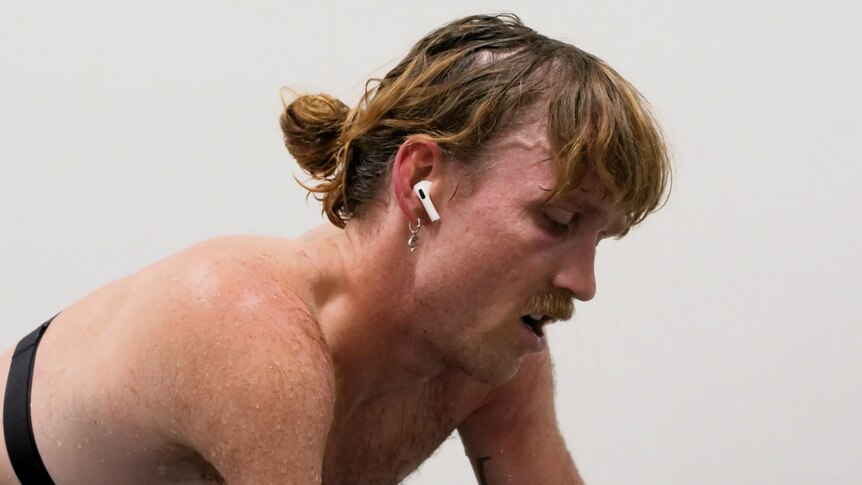 Close up of para cyclist Alistair Donohoe who is shirtless, with air pods, sweating, as he trains in a climate chamber