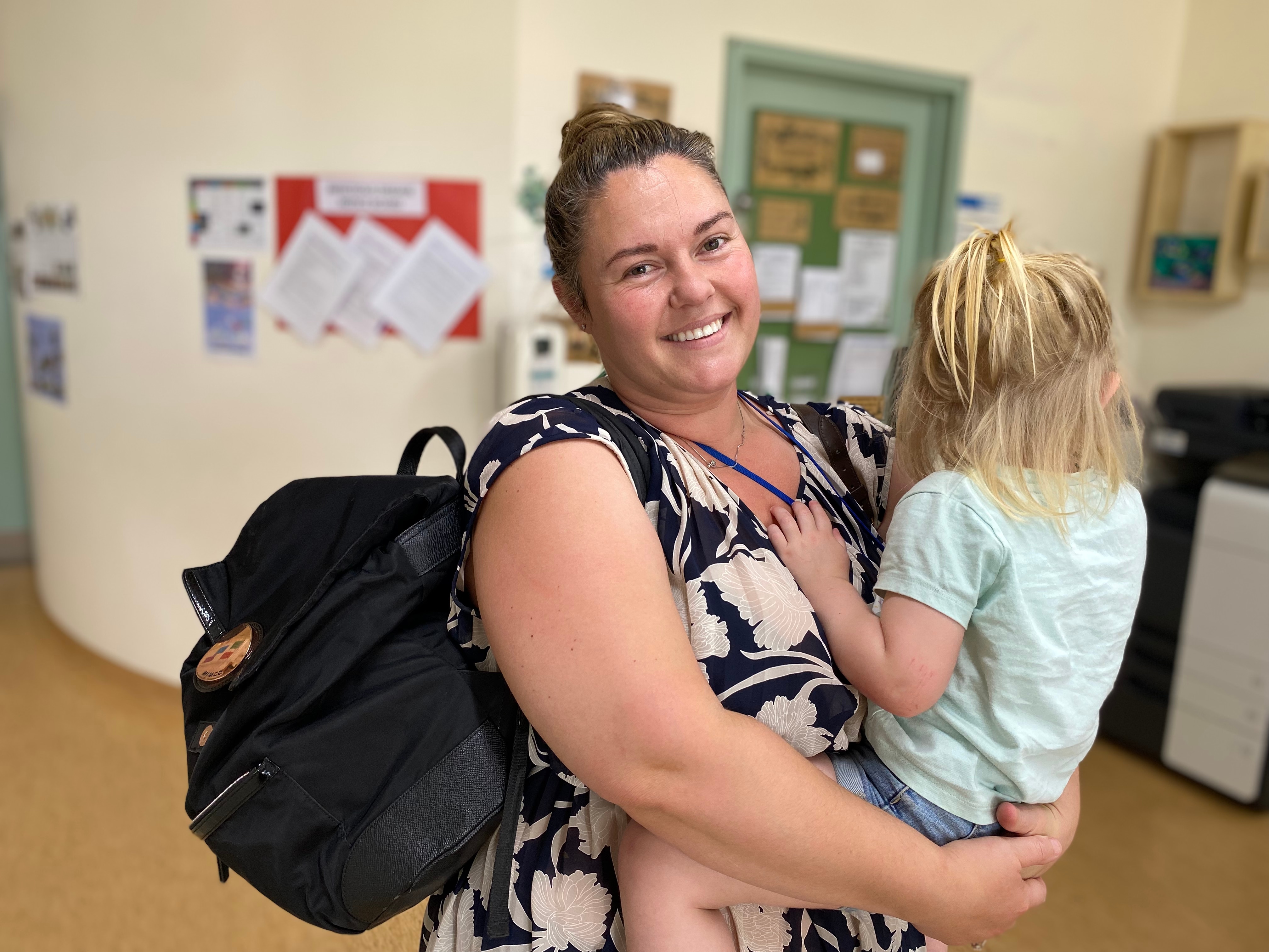 A woman with light brown hair is smiling as she holds a young child wearing a blue shirt with blonde hair.