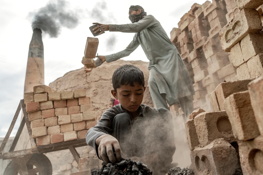 a young boy works on bricks as a person behind lifts bricks on to a pile at a brick factory