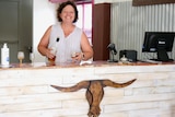 A lady smiling standing behind a bar, is holding a wine bottle and about to pour it in a glass. 