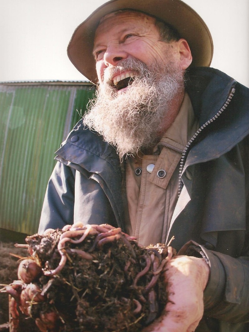 A man with a thick white beard laughs and holds a bundle of soil and earthworms in his hands.