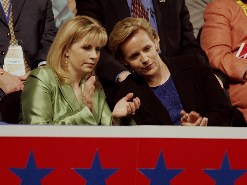 A young Liz Cheney, in a green satin suit, claps while leaning in to talk with Mary Cheney, in a black blazer and blue top