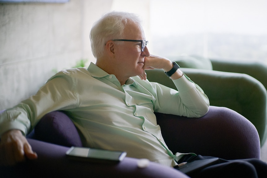 Steve Martin sits on a couch resting his hand on his chin as he stares into the distance on a grey day.