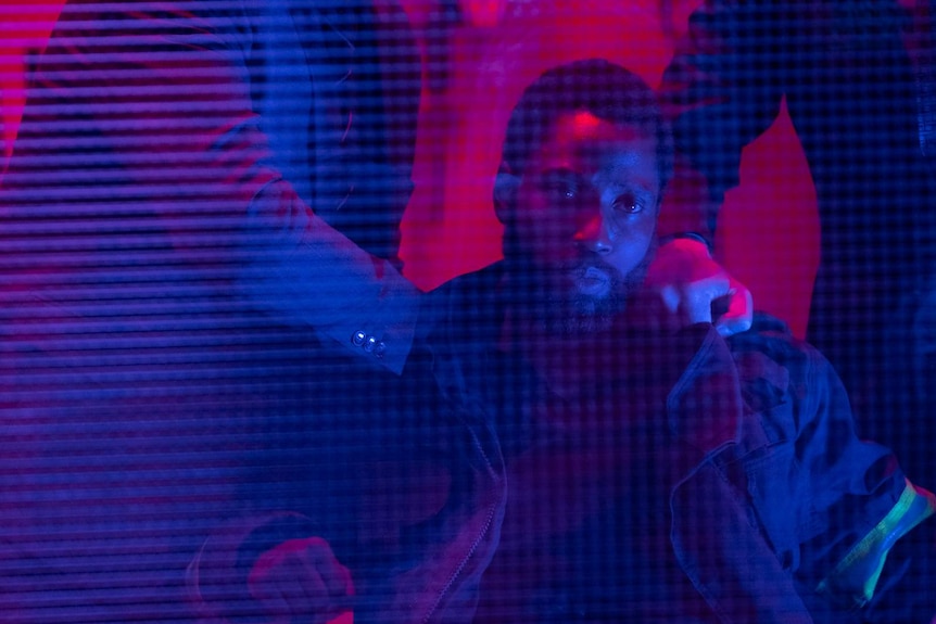 Close-up of actor John David Washington seated, with two sets of hands holding him down, under pink-blue lighting.