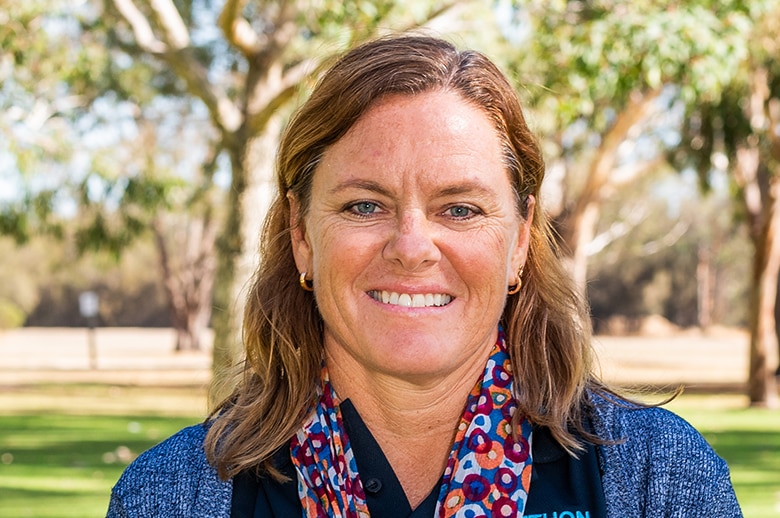 A woman with sandy coloured hair and a bright scarf smiles with trees behind her