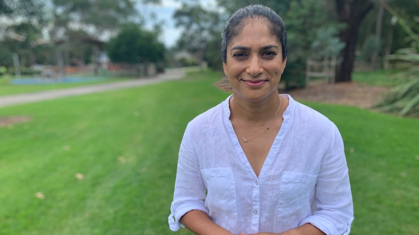 A woman stands in a garden with her hands clasped, looking straight at the camera.