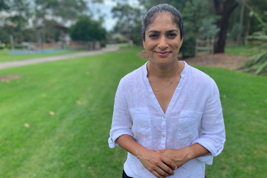 A woman stands in a garden with her hands clasped, looking straight at the camera.