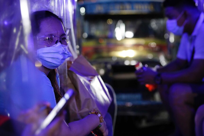 An elderly woman wearing a face mask and glasses sits at night with a bus and masked man behind her.