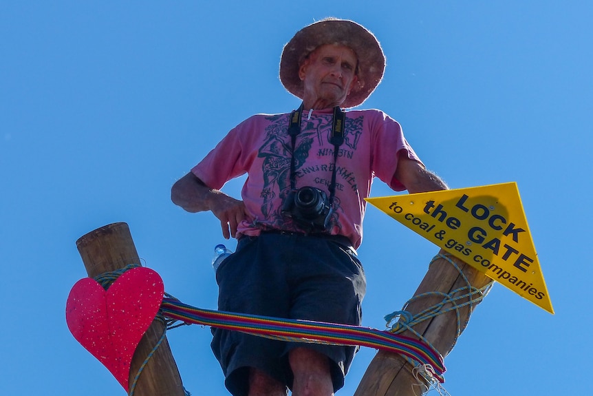 A man in a Nimbin environmental centre short stands atop a wooden post with an anti-CSG sign nearby.