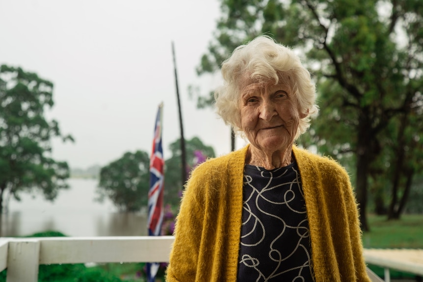 A woman in a yellow cardigan smiles with flood waters behind her.