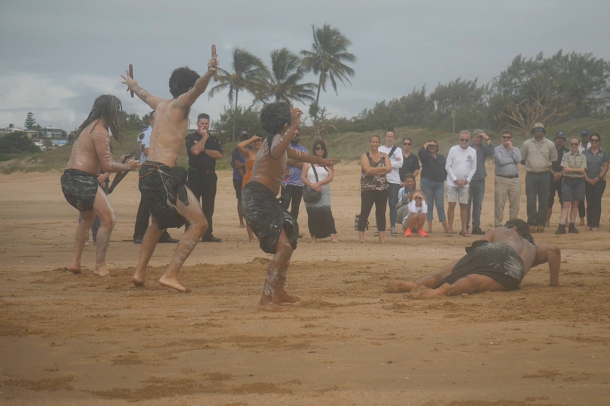 Four dancers in traditional black cloth and white paint dance in sand, people stand watching them. 