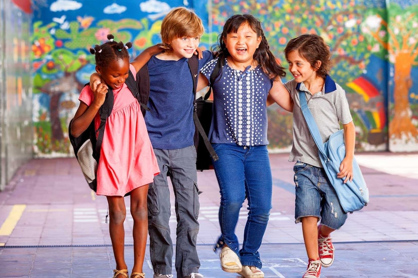 Four young, smiling children with diverse backgrounds in a school courtyard with their arms around one another's shoulders.