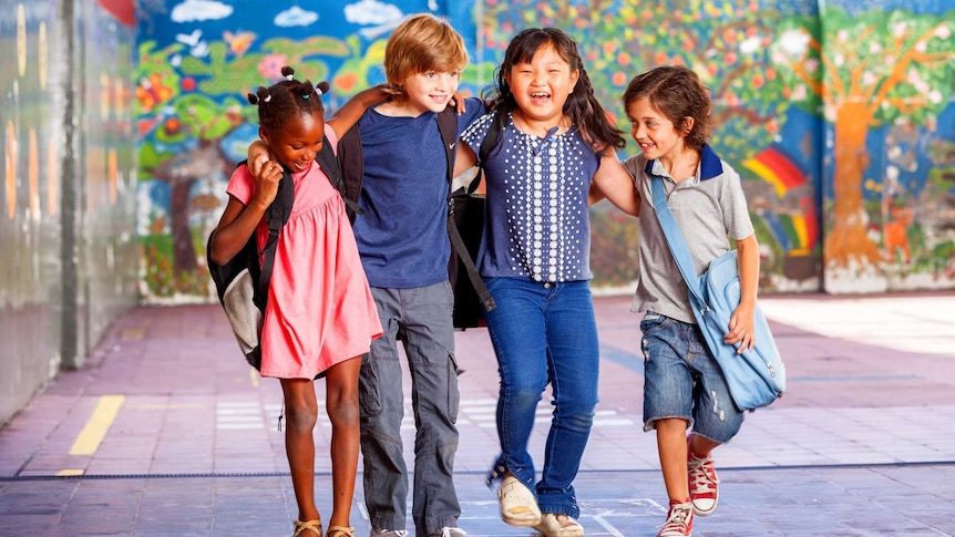 Four young, smiling children with diverse backgrounds in a school courtyard with their arms around one another's shoulders.