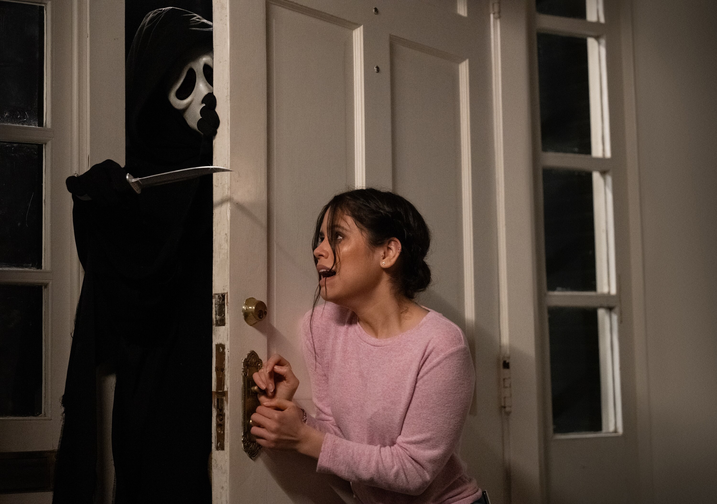 A teenage girl screams as she tries to close the front door on a man wearing a ghost mask and a cape who is wielding a knife