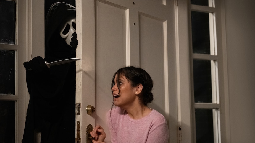 A teenage girl screams as she tries to close the front door on a man wearing a ghost mask and a cape who is wielding a knife