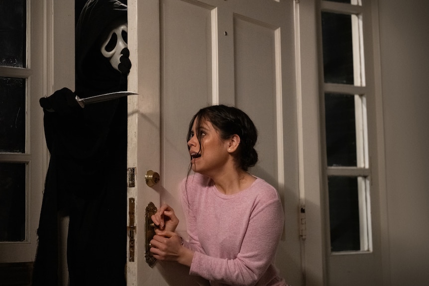 A teenage girl screams as she tries to close the front door on a man wearing a ghost mask and a cape who is wielding a knife