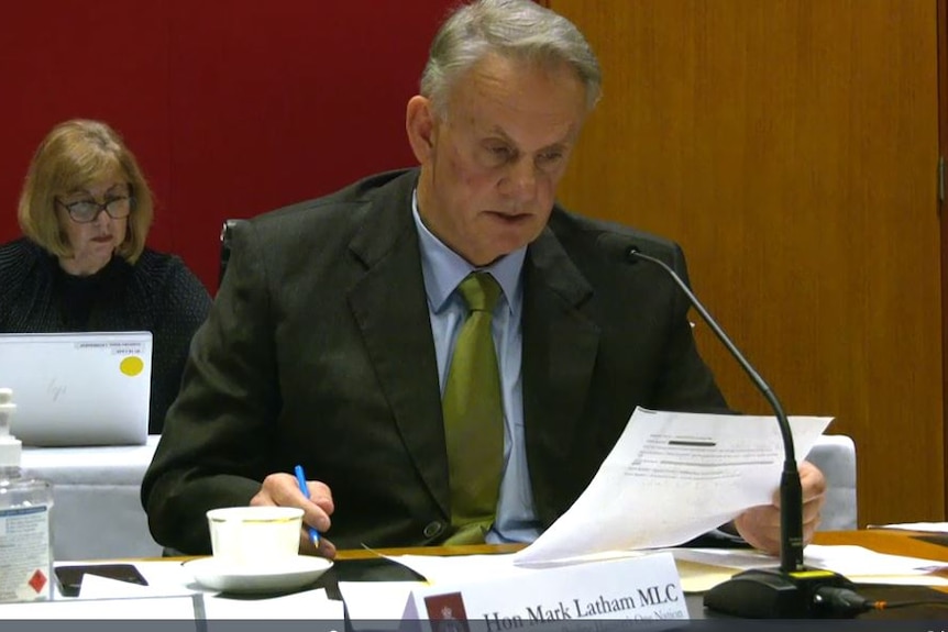 A grey-haired man in a suit – Mark Latham – refers to a document in a parliamentary chamber.