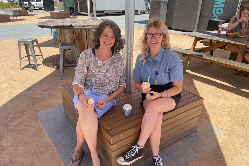 A woman smiles as she sits next to a younger woman on a timber bench seat.