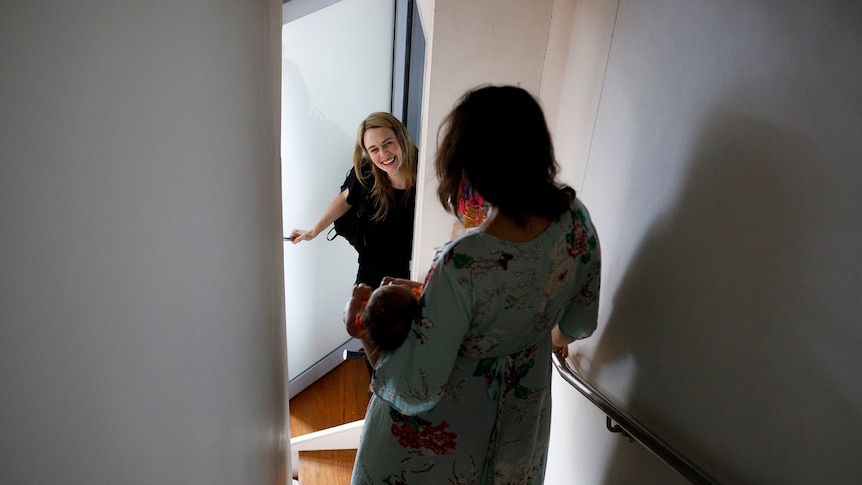 A woman holds a baby in a stairwell while another woman stands in the doorway for a story on share housing.
