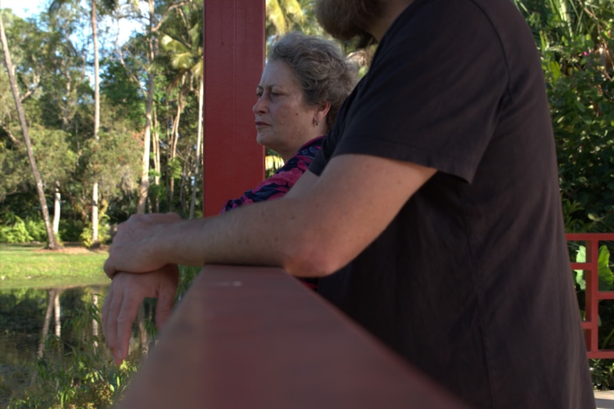 A man's forearms leaning on railing, looking out at a small lake with woman's face in shot.