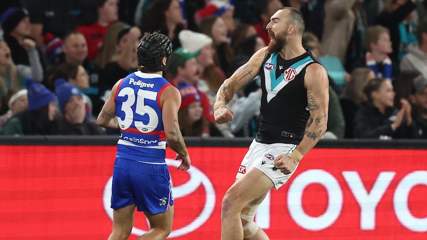 A tall full-forward for Port Adelaide yells in triumph as he celebrates a goal while a defender stands by.
