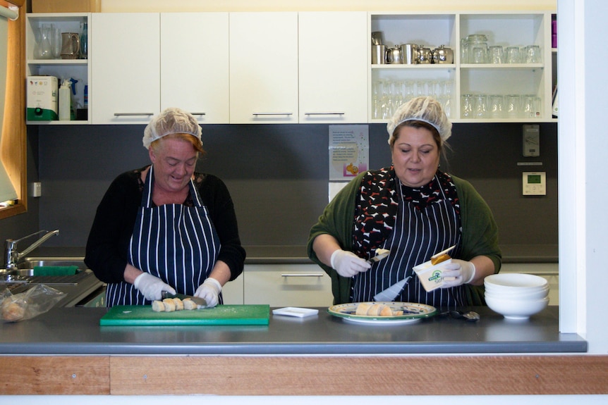 Two women wearing hairnets work in the kitchen of a community house.