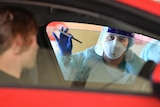 A person in bioprotective gear stands at a passenger side window holding a swab test.