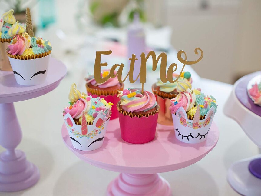 A pink cake stand holds rainbow coloured cupcakes, some with unicorn holders and a sign that reads 'eat me'