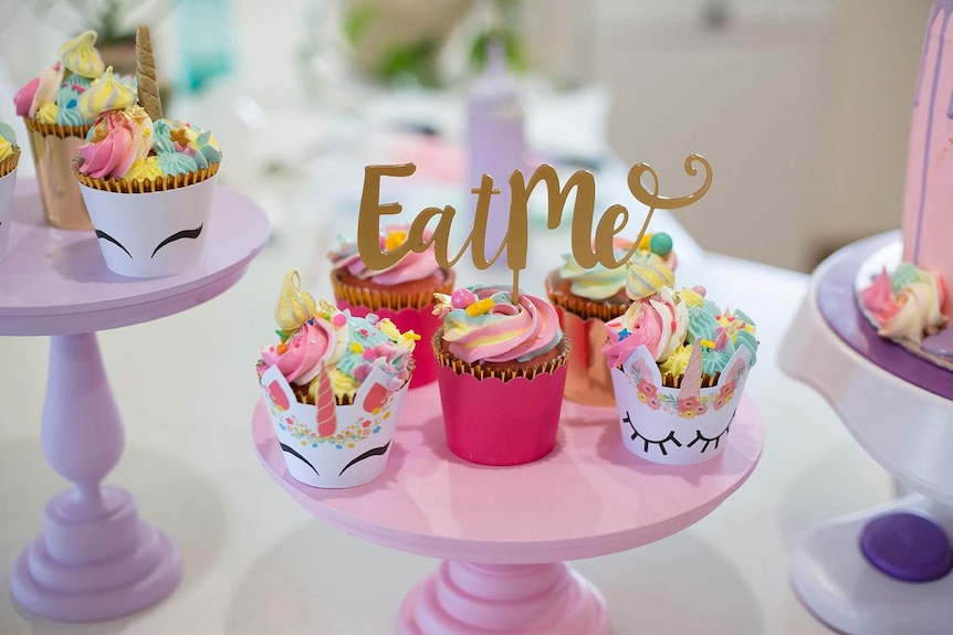 A pink cake stand holds rainbow coloured cupcakes, some with unicorn holders and a sign that reads 'eat me'