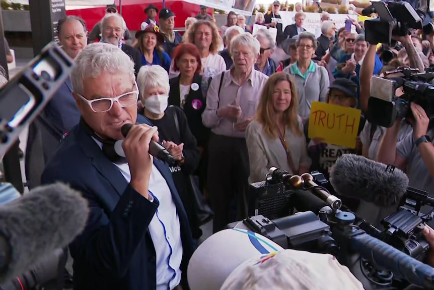 A man in a suit and white-rimmed glasses holds a microphone, surrounded by camera operators and a large crowd.