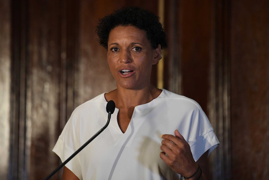 Mel Jones raises her left hand as she speaks behind a microphone and podium.