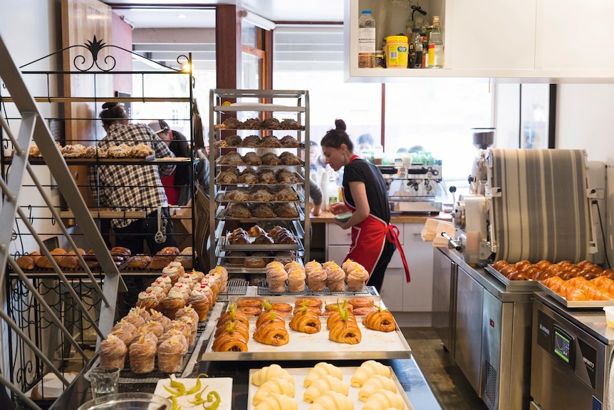 A woman in a red apron putting a tray of croissants in a trolley, many more baked goods surround her
