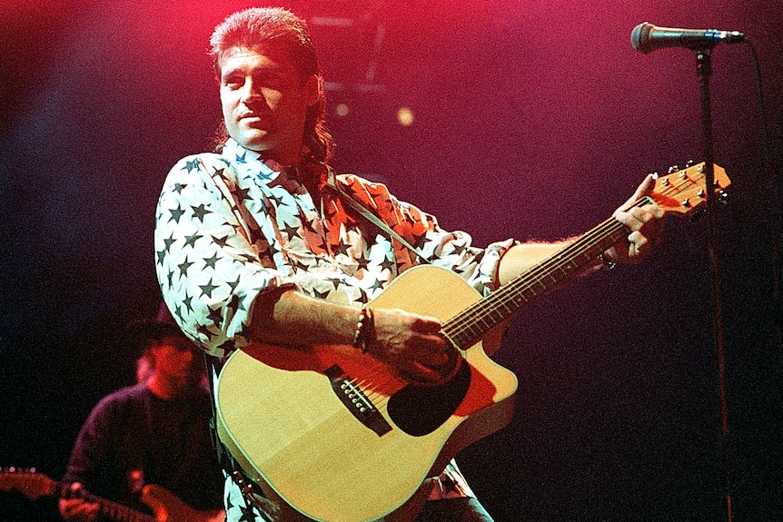 A man with a mullet performs on stage with a guitar. 