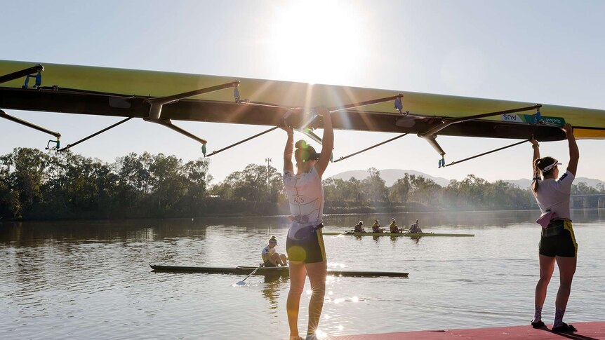 Two women hold a rowing boat above their heads as they prepare to place it in the water. Other rowers in background on river.