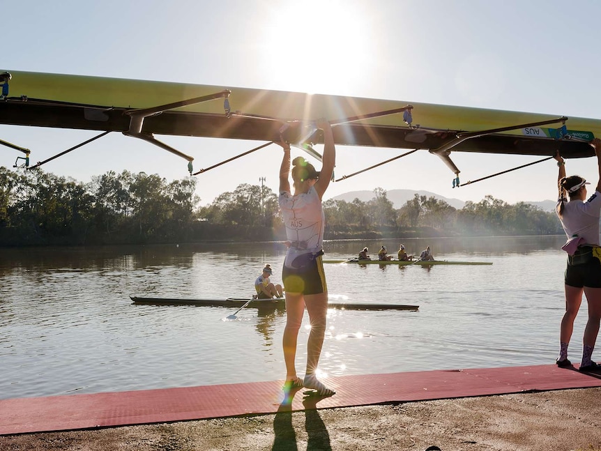 Two women hold a rowing boat above their heads as they prepare to place it in the water. Other rowers in background on river.