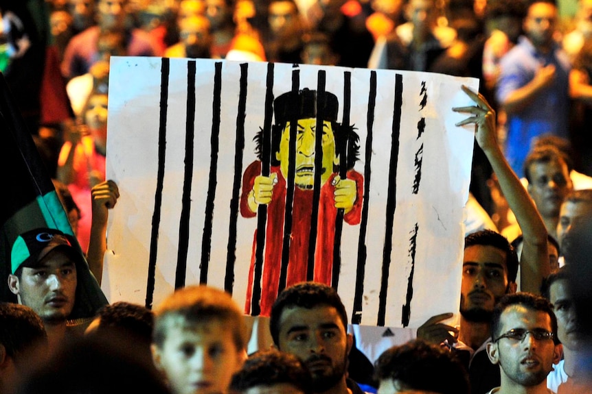 A cartoon of Moamar Gaddafi behind bars is held up in a crowd in Banghazi