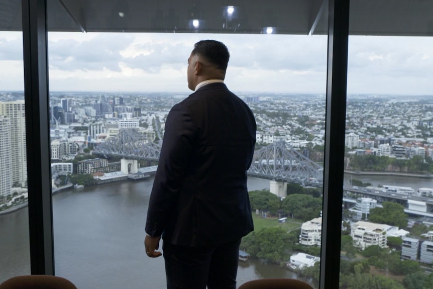 Man in dark suit stands by large boardroom windows overlooking Brisbane river and city. 