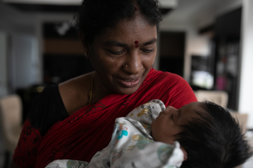 An Indian  woman in black and red saree, black hair, nose ring, bindi, gazes at a newborn baby in her arms. 