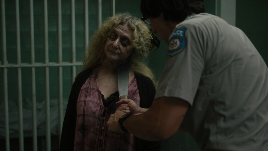 A tall male police officer holds a large knife to the neck of a distressed looking female zombie in a jail cell.
