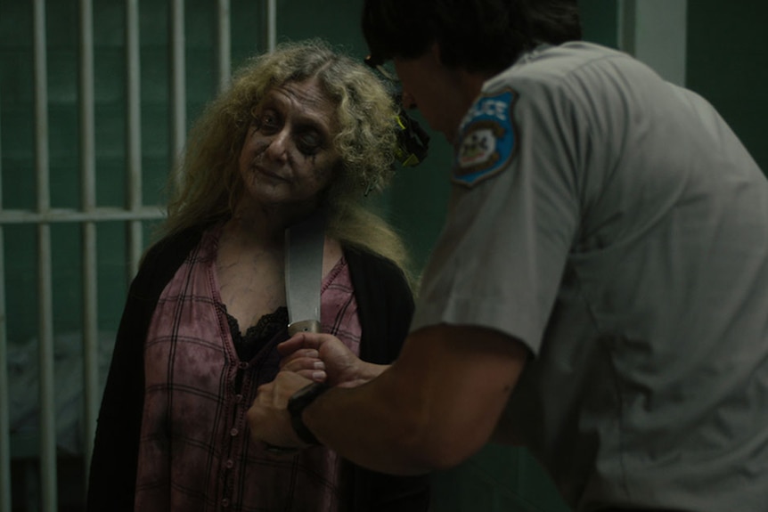 A tall male police officer holds a large knife to the neck of a distressed looking female zombie in a jail cell.