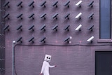 A person wearing all white and a mask with an emoji-style face walks past a wall with dozens of CCTV cameras on it.