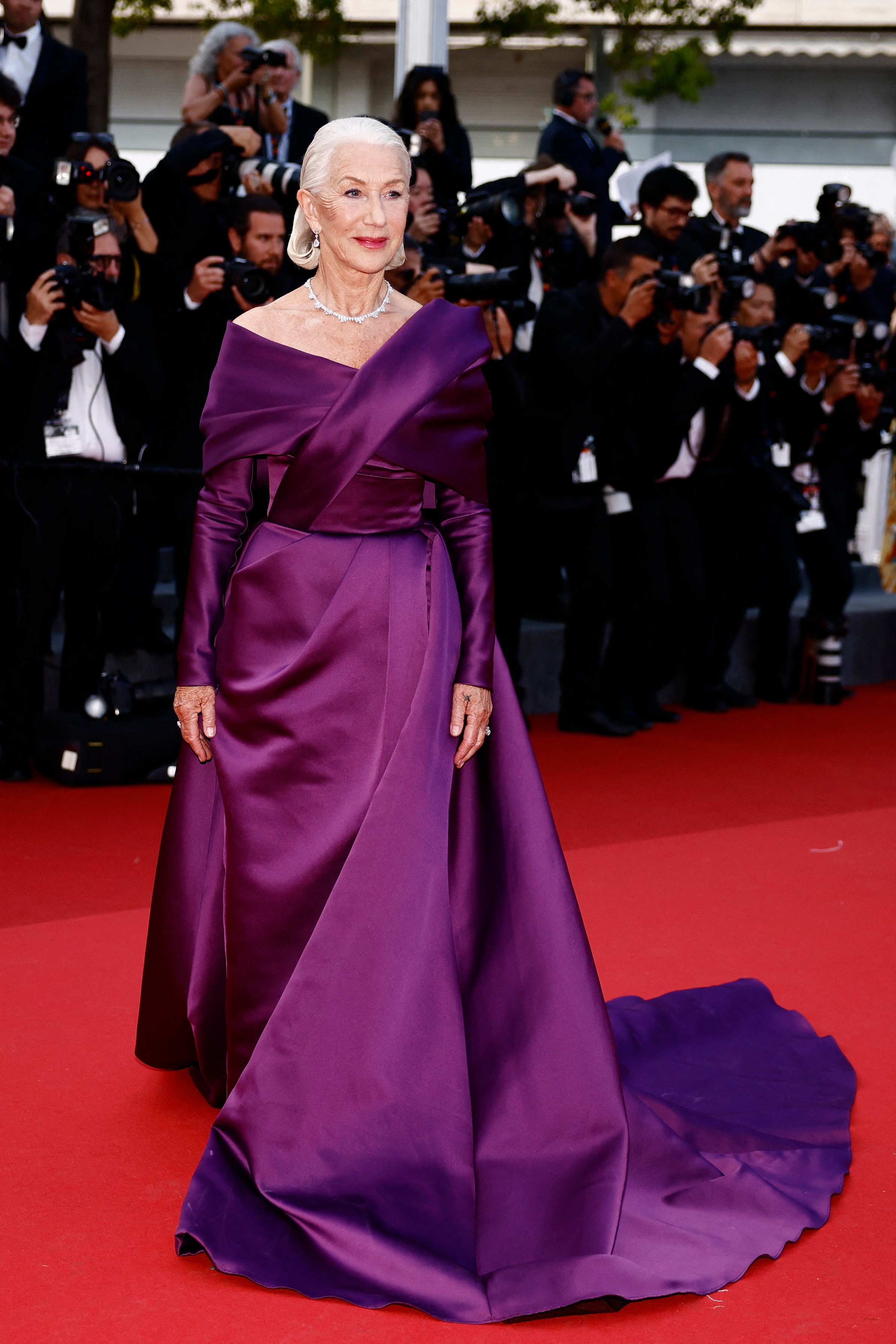 Helen Mirren con un vestido largo y sedoso de color violeta, mangas largas con hombros descubiertos y una ligera cola.