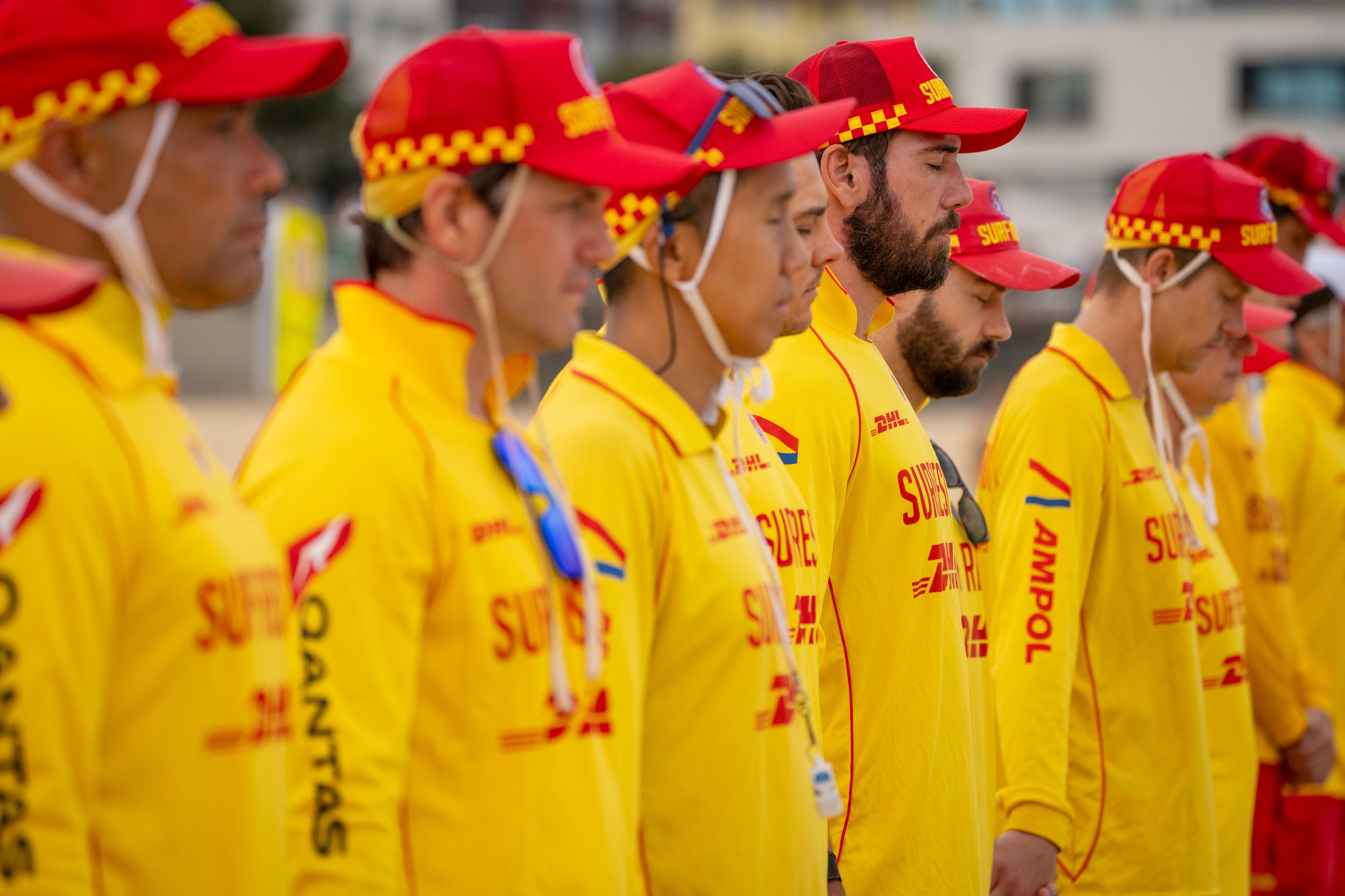 Salva-vidas em uniformes vermelhos e amarelos ficam em fila, alguns fechando os olhos ou baixando a cabeça em um momento de silêncio.