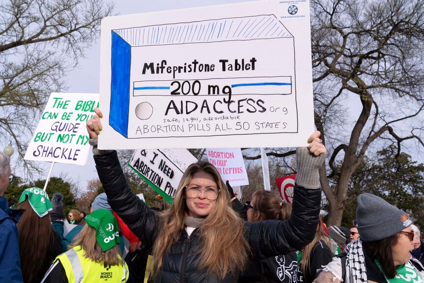At a protest, a woman holds a sign with a drawing of a box of Mifepristone tablets and pro-access slogans.
