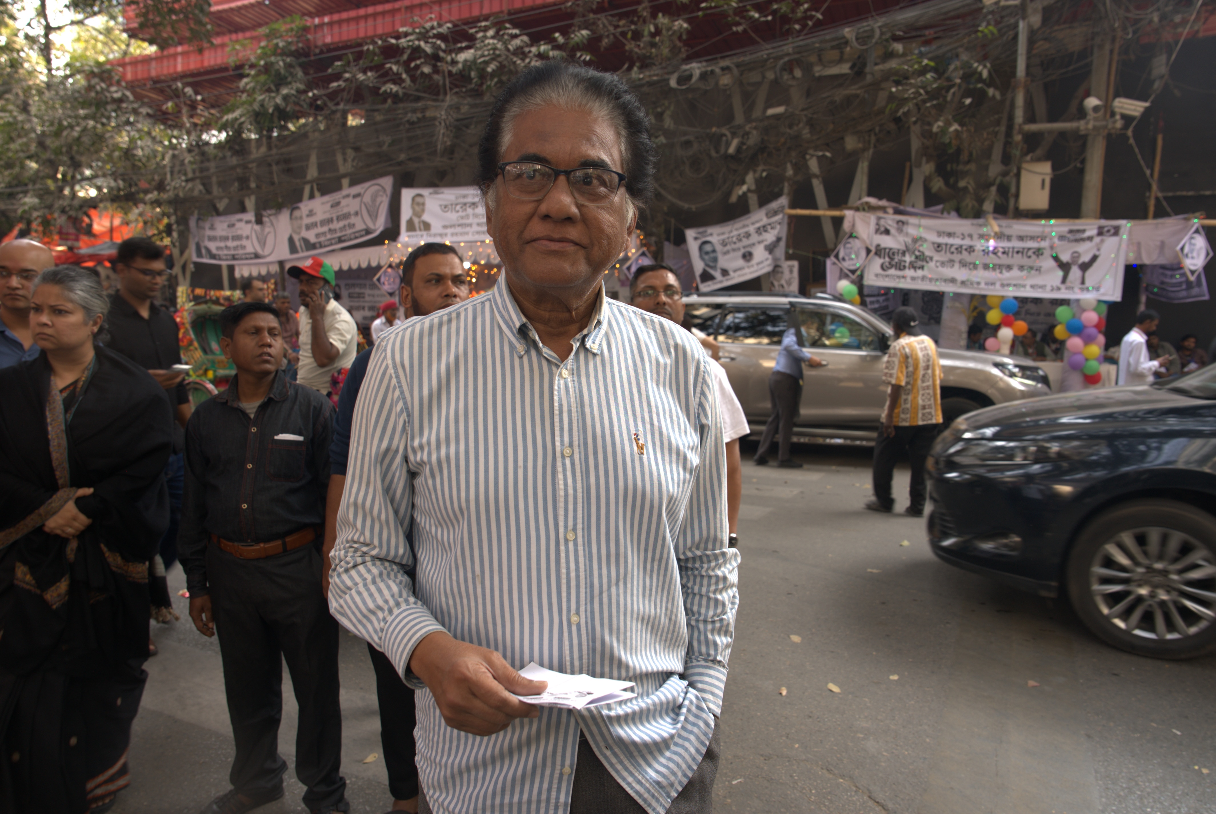 Un hombre con una camisa blanca de cuello a rayas se encuentra en una calle muy transitada y sostiene un trozo de papel en la mano