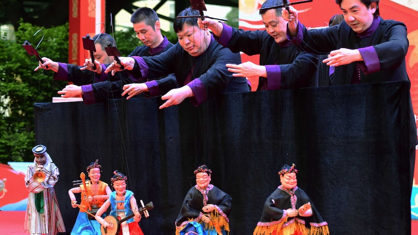 men in black robes with purple shirts underneath dangle string puppets. Outdoor setting