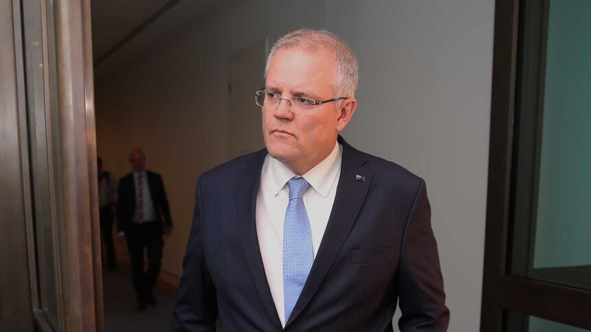 Scott Morrison walks through a doorway. He has one hand in his suit pocket while the other holds a rolled up folder.