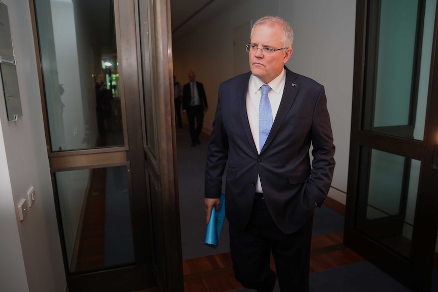 Scott Morrison walks through a doorway. He has one hand in his suit pocket while the other holds a rolled up folder.