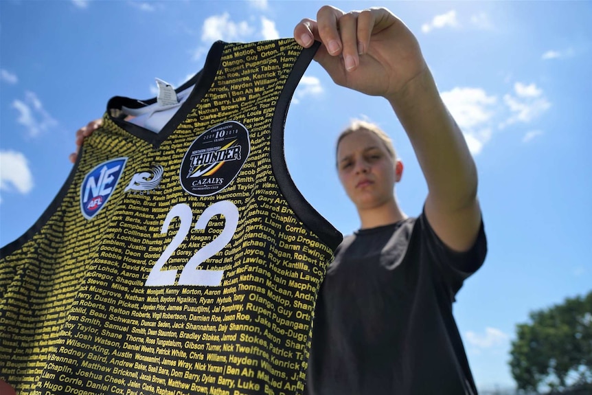 Danielle Ponter holds up and NT Thunder jersey in front of her body with sky and clouds in the background.