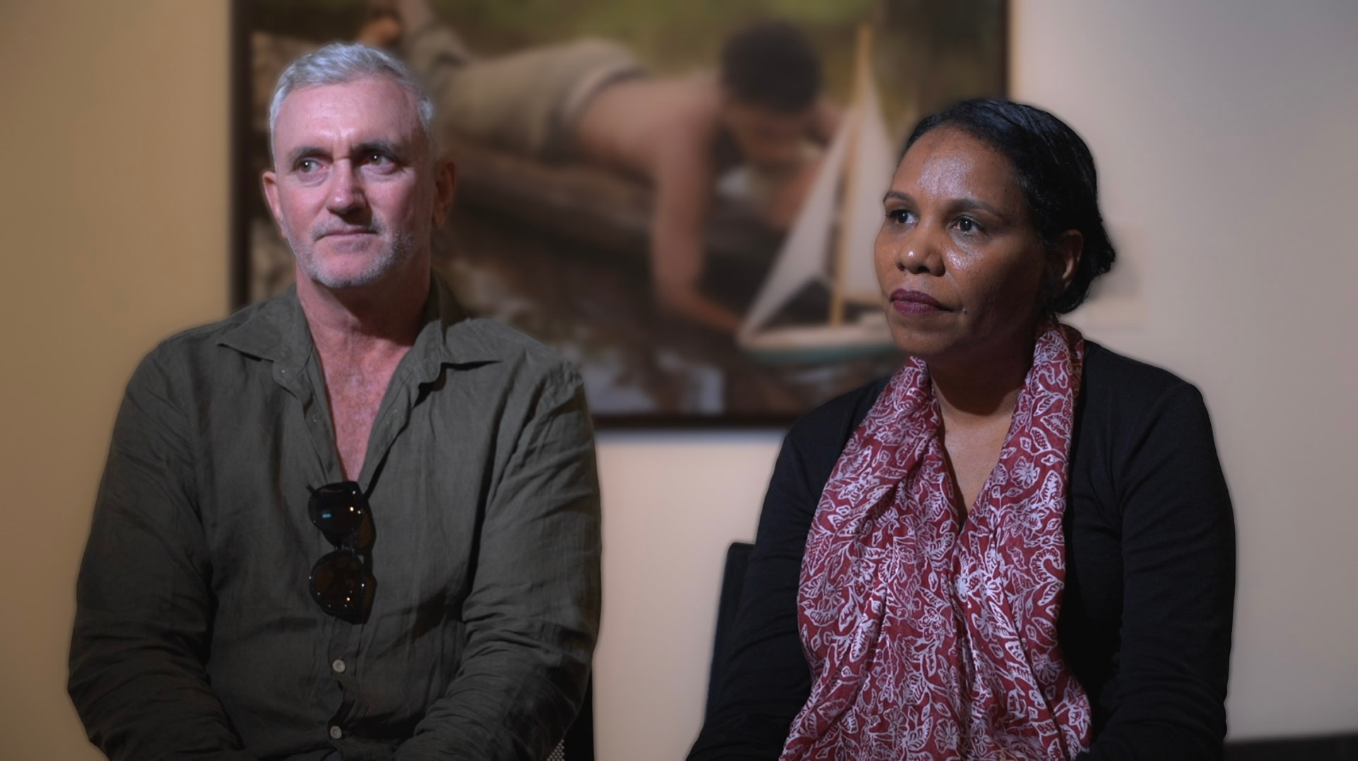A middle-aged man with grey hair sits next to a dark-haired woman in a softly-lit room with a painting on the wall.